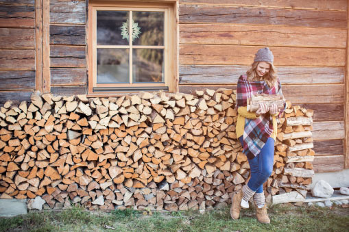 Woman next to a stack of firewood and carrying some to a fire place.